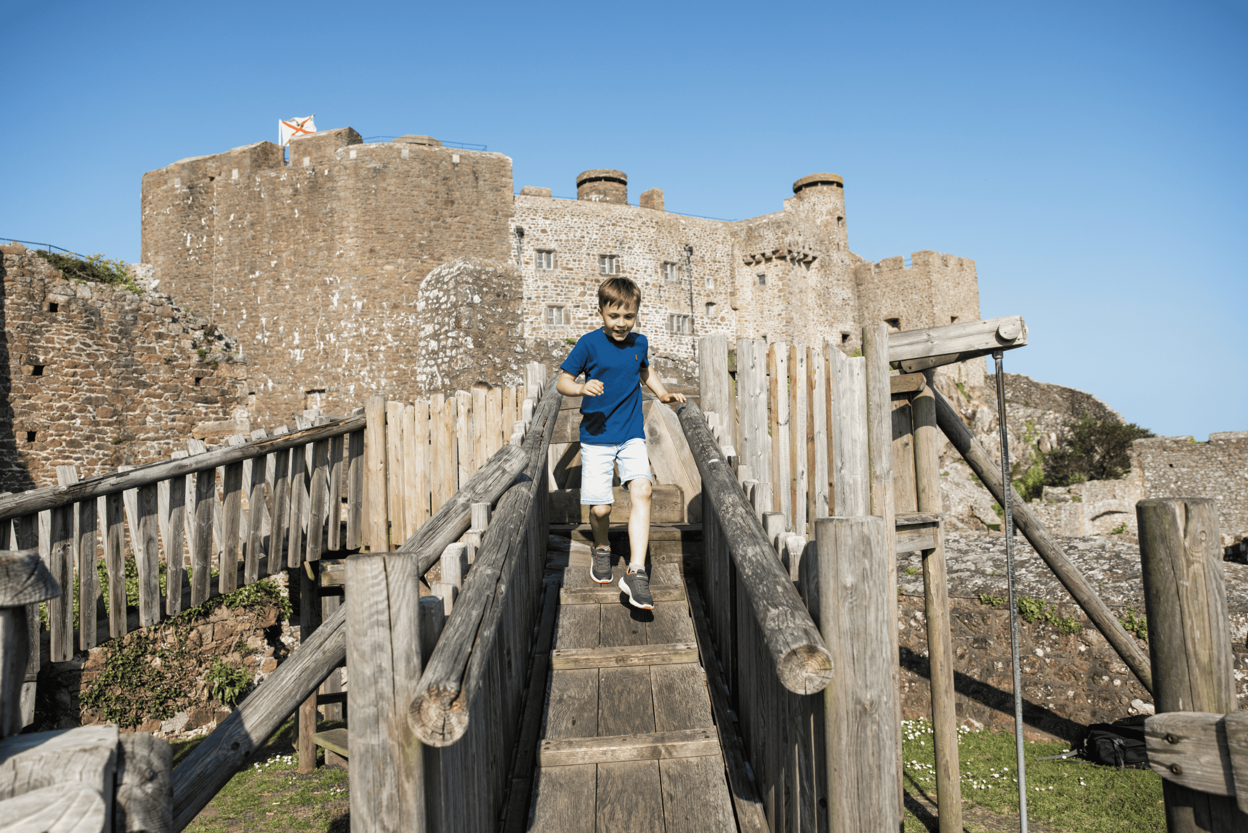 Historic Jersey castle with a child crossing a wooden bridge in front.