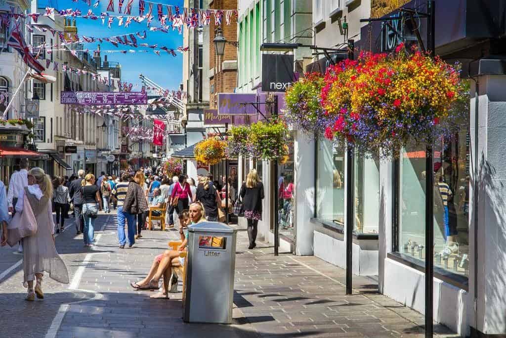 Colourful street scene with pedestrians, shops, and floral decorations.